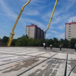 Wylewanie betonu na prefabrykowane płyty stropowe z użyciem pompy do betonu, w tle bloki mieszkalne i błękitne niebo.