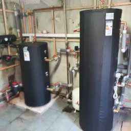 Dwa czarne, cylindryczne zbiorniki, prawdopodobnie na wodę, połączone rurami i zaworami w surowym wnętrzu z drewnianą konstrukcją na ścianach i płytkami na podłodze. Na jednym zbiorniku widoczna...