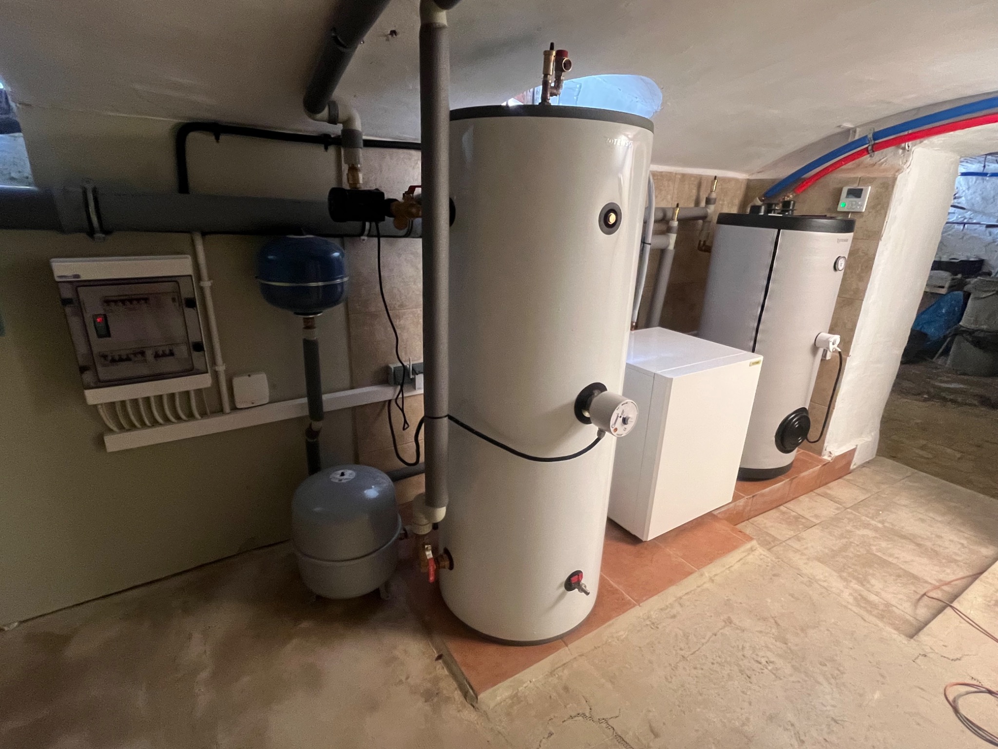 Dwa białe bojlery w piwnicy, z widoczną instalacją hydrauliczną, skrzynką elektryczną i rurami grzewczymi pod sufitem.