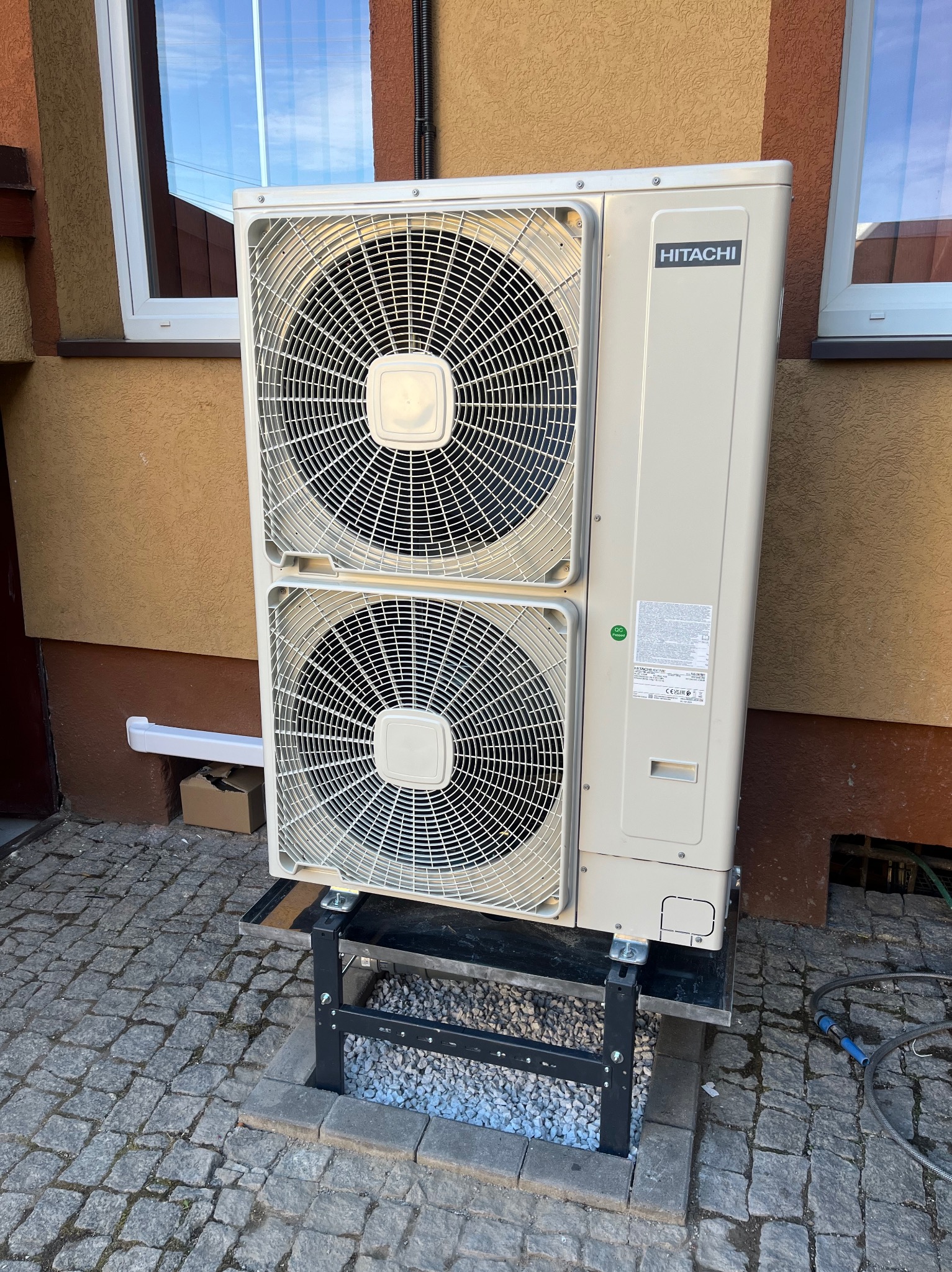 Zewnętrzna jednostka klimatyzacji marki Hitachi, zamontowana na metalowej konstrukcji na kostce brukowej, widok na elewację budynku z dwoma oknami.