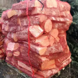 Paleta drewna kominkowego w czerwonej siatce, widoczne końcówki polan z korą, na tle iglastego drzewa.