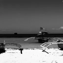 Czarno-białe zdjęcie trzech tradycyjnych filipińskich łodzi banca na piaszczystej plaży, z widocznym horyzontem morskim i odległym statkiem.