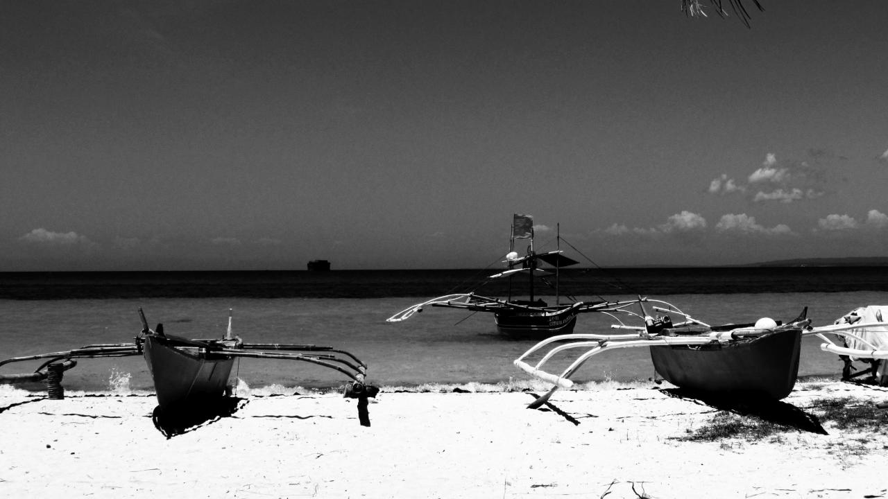Czarno-białe zdjęcie trzech tradycyjnych filipińskich łodzi banca na piaszczystej plaży, z widocznym horyzontem morskim i odległym statkiem.