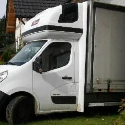 Biały samochód dostawczy Renault Master z wysoką kabiną i plandeką, zaparkowany na trawie przed domem z czarnym dachem, prawe lusterko owinięte czarną folią.