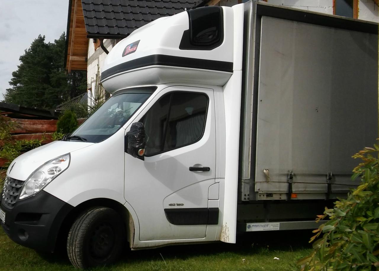 Biały samochód dostawczy Renault Master z wysoką kabiną i plandeką, zaparkowany na trawie przed domem z czarnym dachem, prawe lusterko owinięte czarną folią.