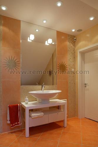 Nowoczesna łazienka z terakotową podłogą i ścianami w odcieniach pomarańczy, biała umywalka nablatowa na szafce, duże lustro z oświetleniem punktowym, dekoracyjne motywy słońca na ścianach.