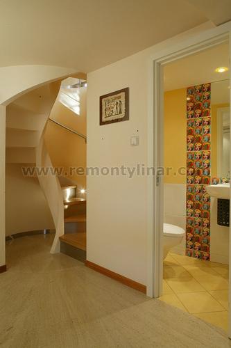 Wnętrze domu z widokiem na spiralne schody z podświetleniem oraz otwarte drzwi do żółtej łazienki z wzorzystą mozaiką i białą armaturą.