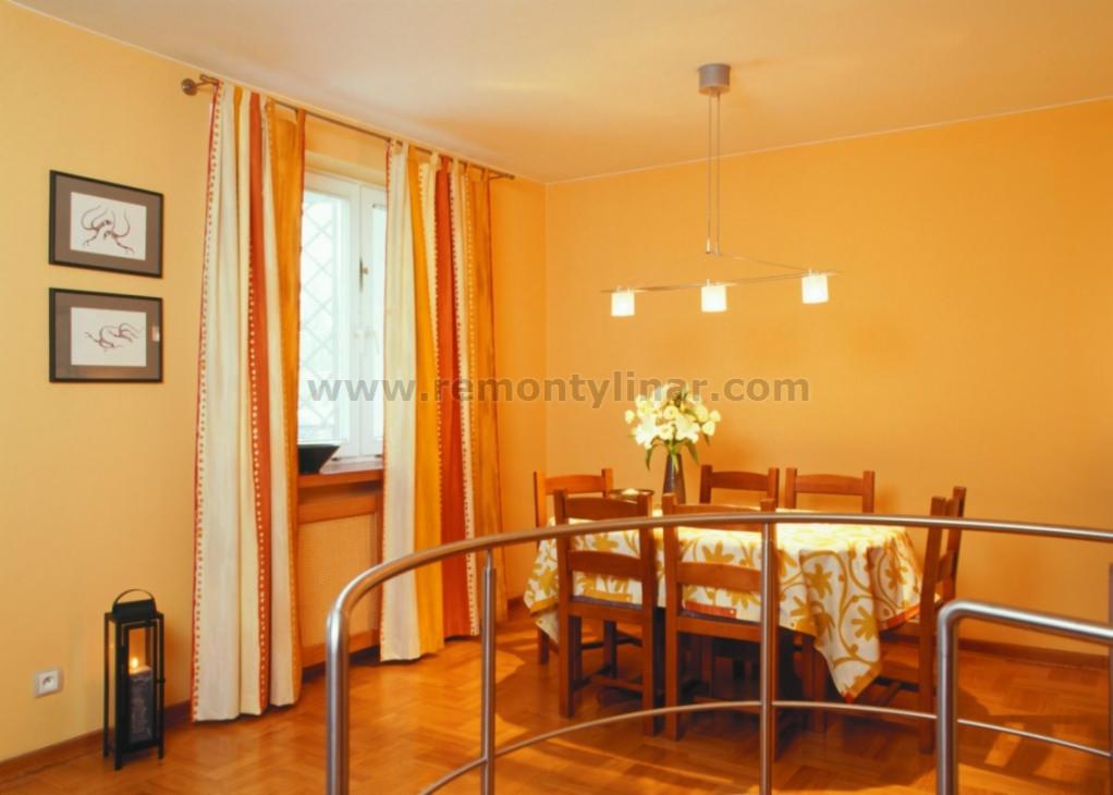 Jasna jadalnia z pomarańczowymi ścianami, drewnianym parkietem i stołem nakrytym wzorzystym obrusem, widok zza metalowej barierki.