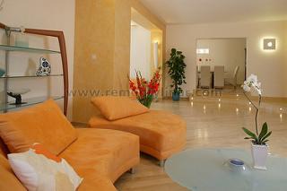 Jasne wnętrze salonu z marmurową podłogą, pomarańczowymi szezlongami i szklanym stolikiem kawowym. W tle widoczna jadalnia i rośliny doniczkowe. Na ścianie kwadratowa lampa.