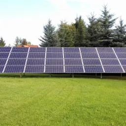 Duża instalacja paneli słonecznych na metalowych podporach na zielonej trawie, w tle wysokie drzewa iglaste i budynek z czerwonym dachem.