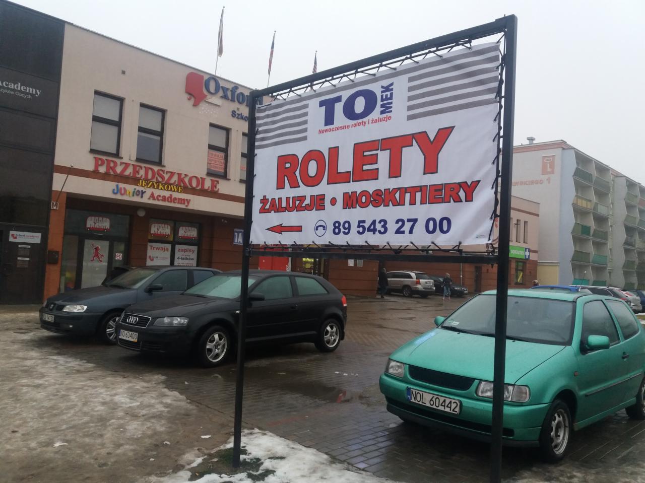 Billboard reklamujący rolety, żaluzje i moskitiery na tle budynku z przedszkolem językowym i zaparkowanymi samochodami, w pochmurny dzień.