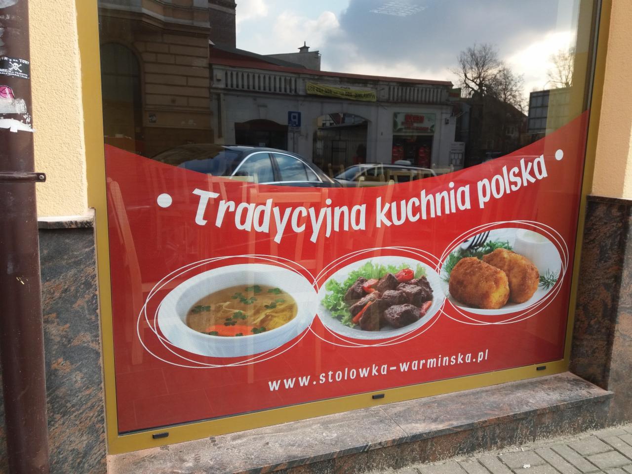 Czerwony baner reklamowy w oknie lokalu gastronomicznego z hasłem 'Tradycyjna kuchnia polska' i zdjęciami zupy, mięsa i kotletów, odbicia ulicy w szybie.