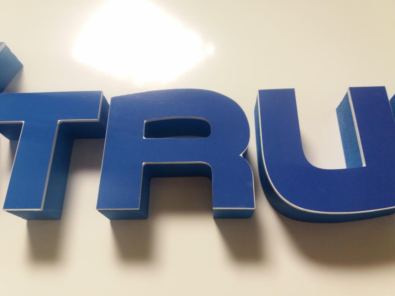 Niebieskie, trójwymiarowe litery z białym obrysem, tworzące fragment słowa, ustawione na białej, błyszczącej powierzchni.