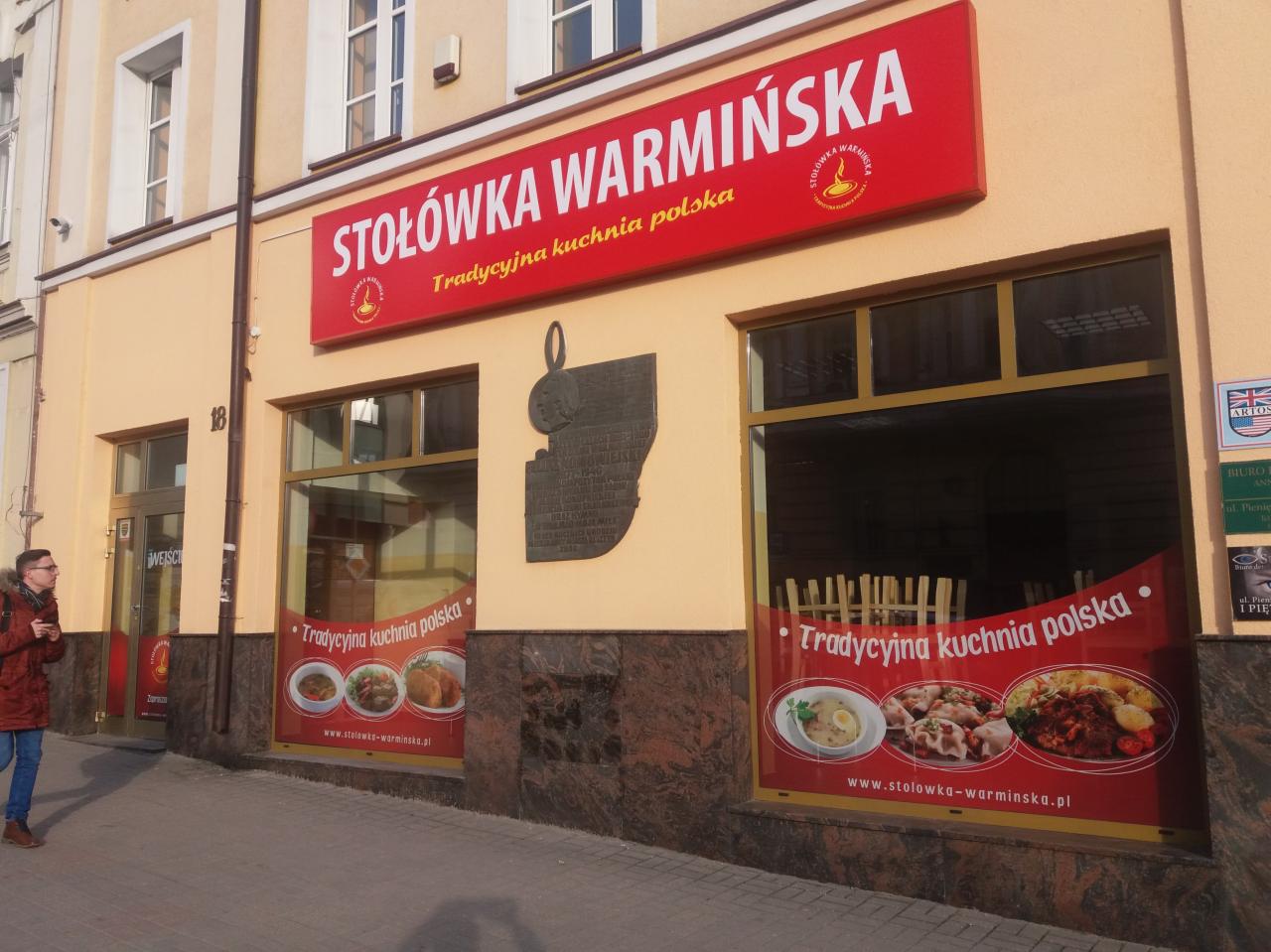 Fasada budynku z szyldem 'Stołówka Warmińska' i oklejonymi witrynami z motywem tradycyjnej kuchni polskiej, widoczny przechodzień w kurtce.