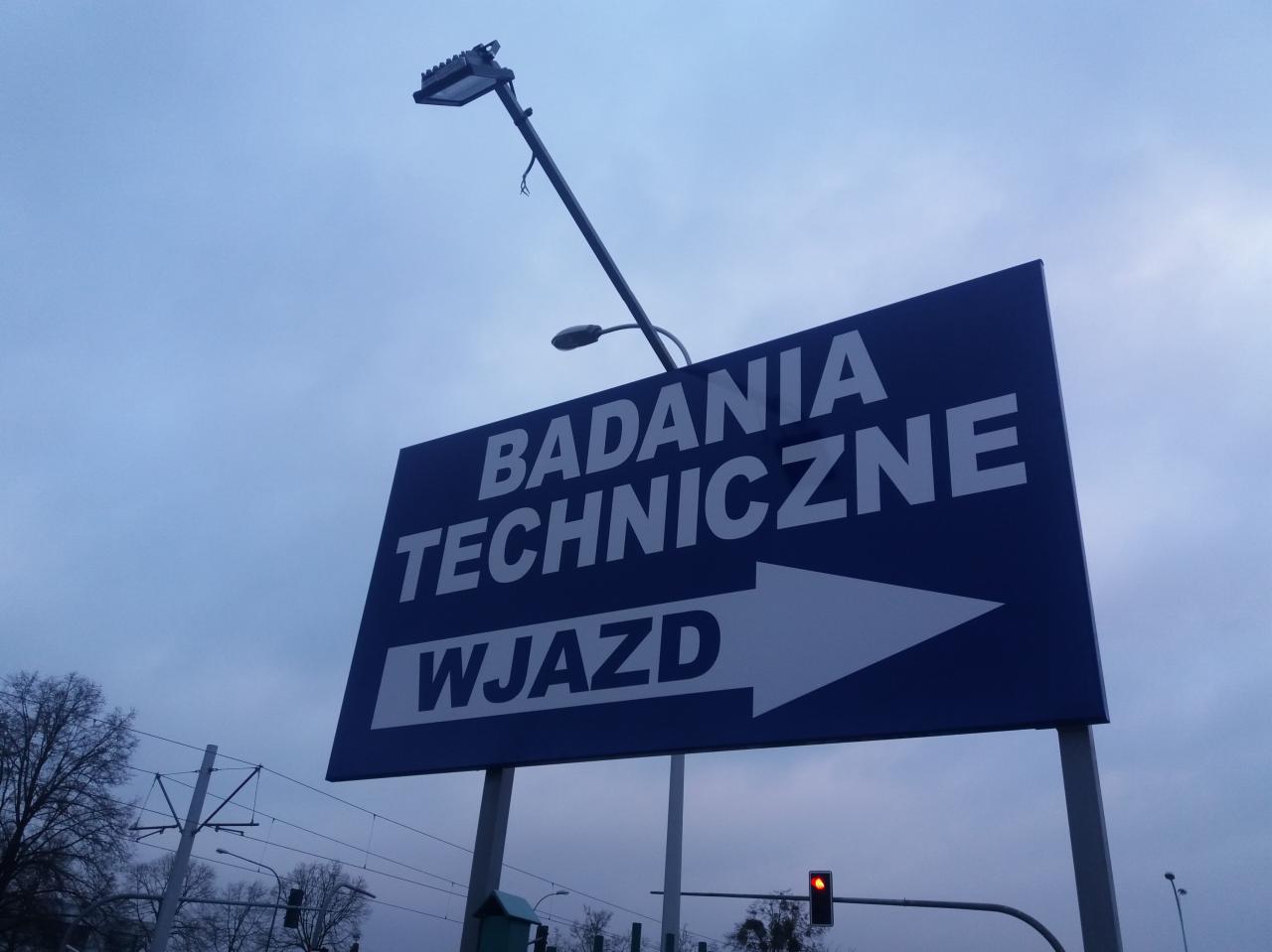 Niebieski szyld informacyjny z napisem 'Badania Techniczne Wjazd' i strzałką, umieszczony na słupie, z widoczną lampą uliczną i sygnalizacją świetlną w tle.