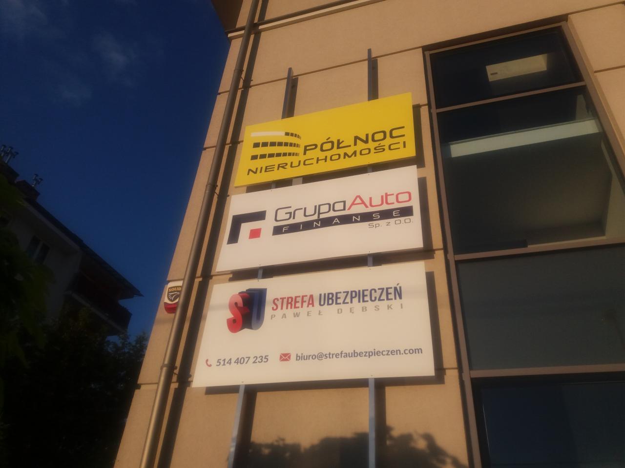 Zewnętrzny szyld reklamowy z logo firm: Północ Nieruchomości, Grupa Auto Finanse i Strefa Ubezpieczeń Paweł Dębski, umieszczony na ścianie budynku biurowego.
