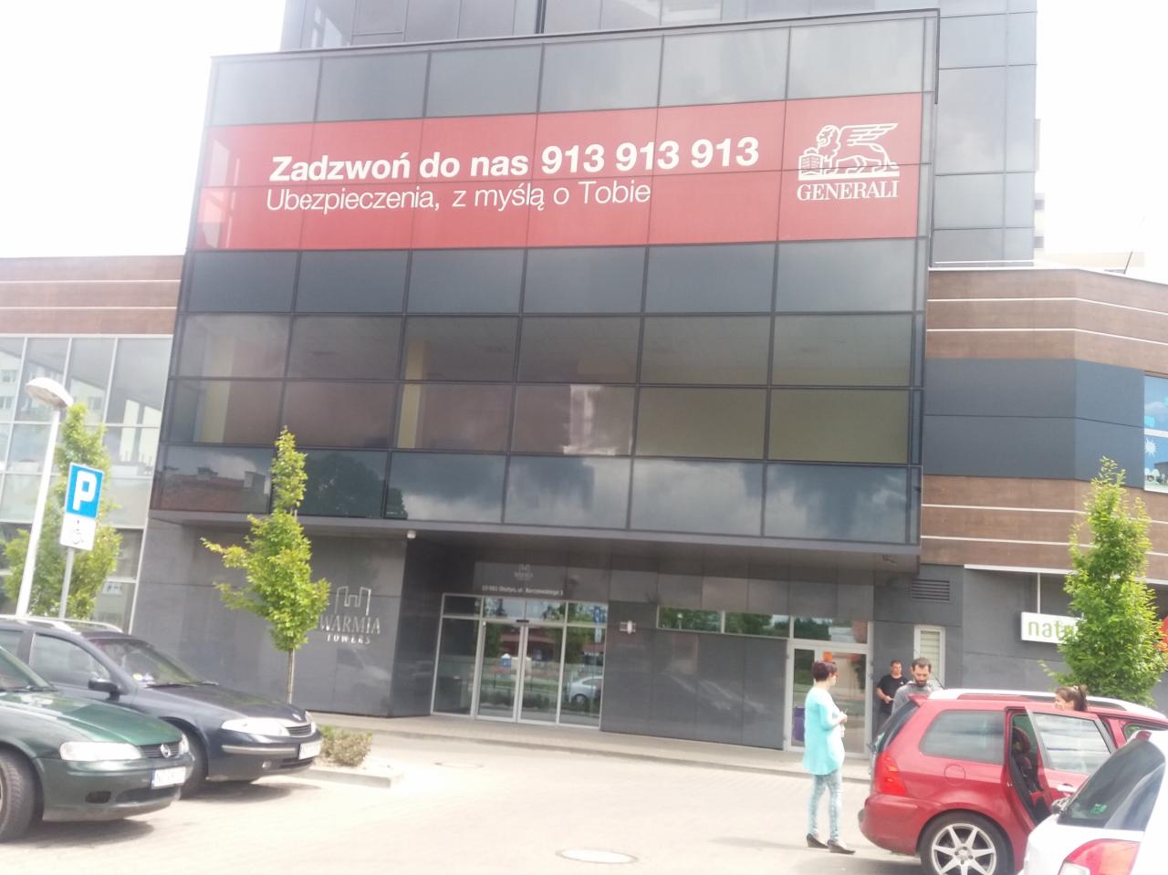 Budynek biurowy z dużą reklamą firmy ubezpieczeniowej Generali, z numerem telefonu i hasłem 'Ubezpieczenia, z myślą o Tobie', widoczny parking i kilka osób w pobliżu wejścia.