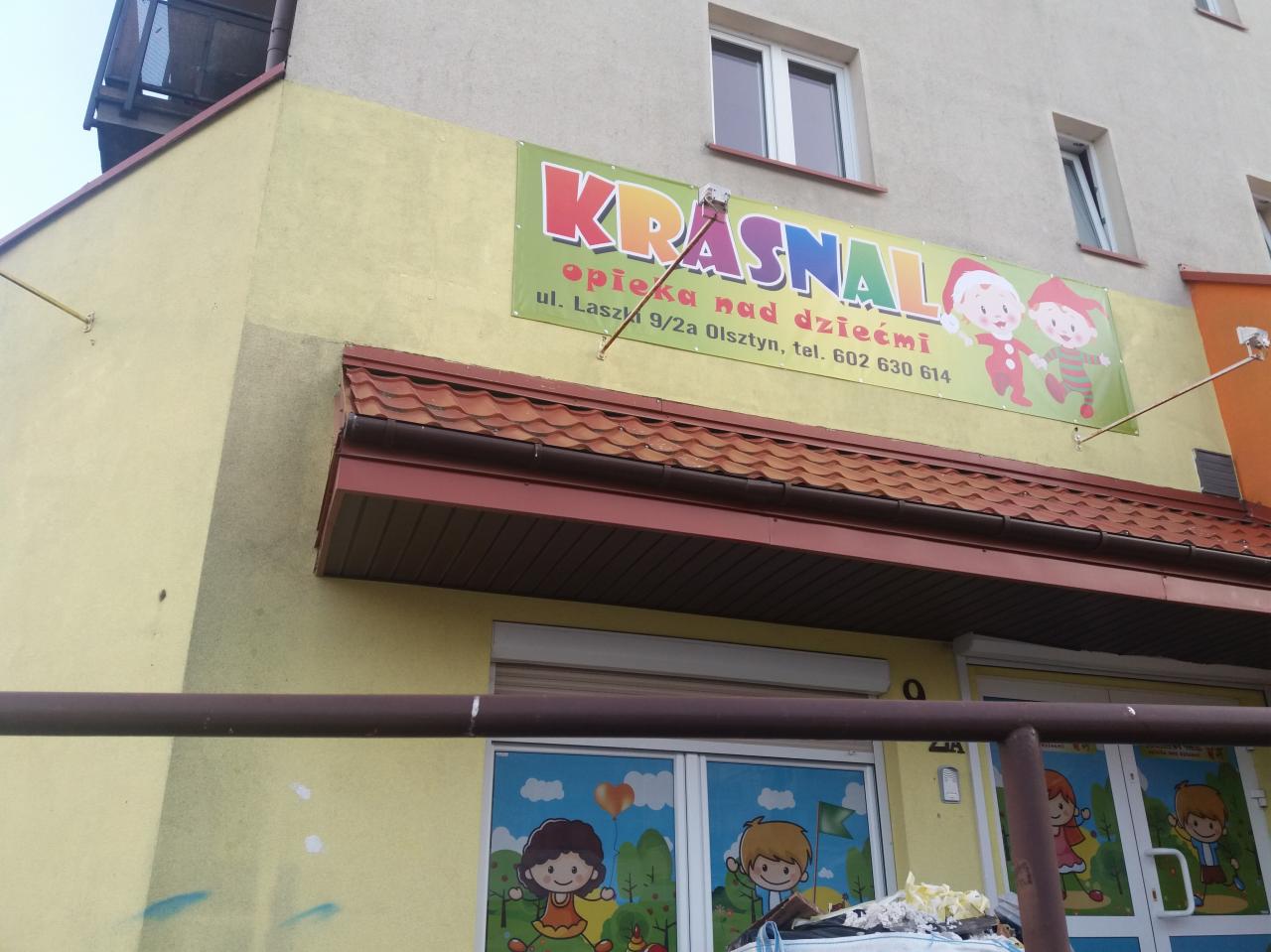 Żółty budynek z zielonym szyldem 'KRASNAL' i rysunkami dzieci, opieka nad dziećmi, adres i telefon w Olsztynie, okna z naklejkami z postaciami z bajek