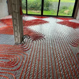 Rozłożone rury ogrzewania podłogowego w surowym wnętrzu, widoczne pętle i mocowania na folii z siatką, w tle okno z widokiem na zieleń.