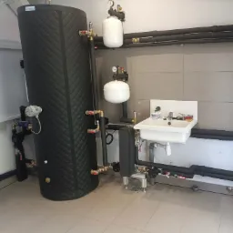 Czarny, ocieplony zbiornik buforowy z osprzętem hydraulicznym, manometrami i naczyniami przeponowymi w kotłowni z umywalką i szarymi płytkami ściennymi.