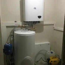 Biały, wiszący piec gazowy Ariston, podłączony do pionowego, białego zasobnika ciepłej wody, z widoczną niebieską przeponą, żółtą pompą REMS i kanistrami, w pomieszczeniu technicznym.