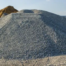 Góra szarego, drobnego kruszywa na placu budowy, z widocznymi śladami opon na ziemi i hałdami piasku w tle.