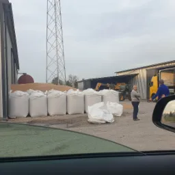 Magazyn zboża: białe worki z ziarnem, koparka JCB, pracownicy i ciężarówki w tle, widok zza szyby samochodu.