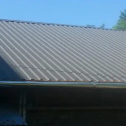 Szary dach z metalu falistego, widoczna nowa, błyszcząca rynna, tło niebieskie niebo i zielone drzewa.