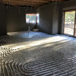 Instalacja ogrzewania podłogowego w surowym wnętrzu, rury grzewcze ułożone na siatce, widoczne okno i drzwi balkonowe, ściany w stanie deweloperskim.