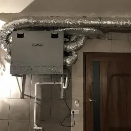 Szara centrala wentylacyjna z logo WindMaker Phantom zamontowana na ścianie z szarych płytek, z podłączonymi rurami wentylacyjnymi i instalacją hydrauliczną, obok drewnianych drzwi.