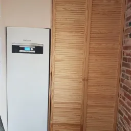 Biała jednostka grzewcza Buderus z cyfrowym wyświetlaczem zamontowana obok drewnianej szafy żaluzjowej w Myszakówku, na tle ściany z czerwonej cegły.