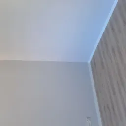 Świeżo wykończone wnętrze: biały sufit z listwą, szara ściana i podłoga z paneli w odcieniach szarości, widoczny fragment gniazdka elektrycznego.