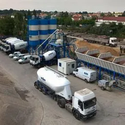 Widok z góry na rozległy teren betoniarni z silosami w biało-niebieskie pasy, rzędem betonomieszarek, zasobnikami na kruszywo i stertami piasku. Na drugim planie zabudowa miejska i drzewa.