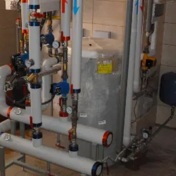 Złożona instalacja kotłowni gazowej z rurami, zaworami, manometrami, pompami obiegowymi i zbiornikiem wyrównawczym, widoczna na tle płytek ceramicznych.