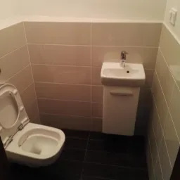 Mała łazienka z beżowymi płytkami na ścianach, ciemną podłogą, wiszącą umywalką z szafką i toaletą z otwartą klapą.