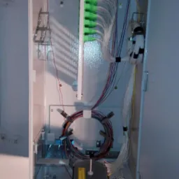 Otwarta skrzynka telekomunikacyjna z widocznymi kablami światłowodowymi, złączkami i elementami montażowymi wewnątrz, zamontowana na ścianie z widocznymi śladami zabrudzeń.