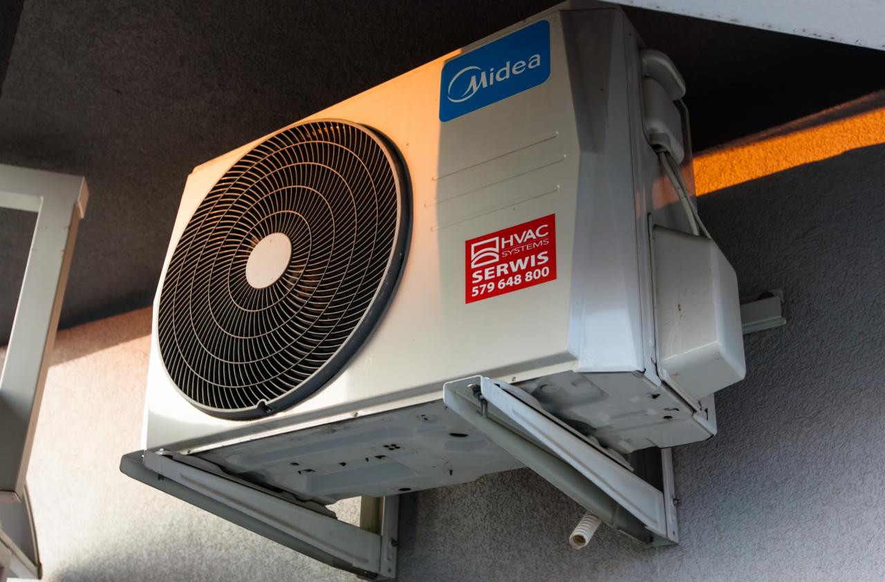 Zewnętrzna jednostka klimatyzacji marki Midea zamontowana na ścianie budynku, z widoczną naklejką serwisową HVAC Systems z numerem telefonu.