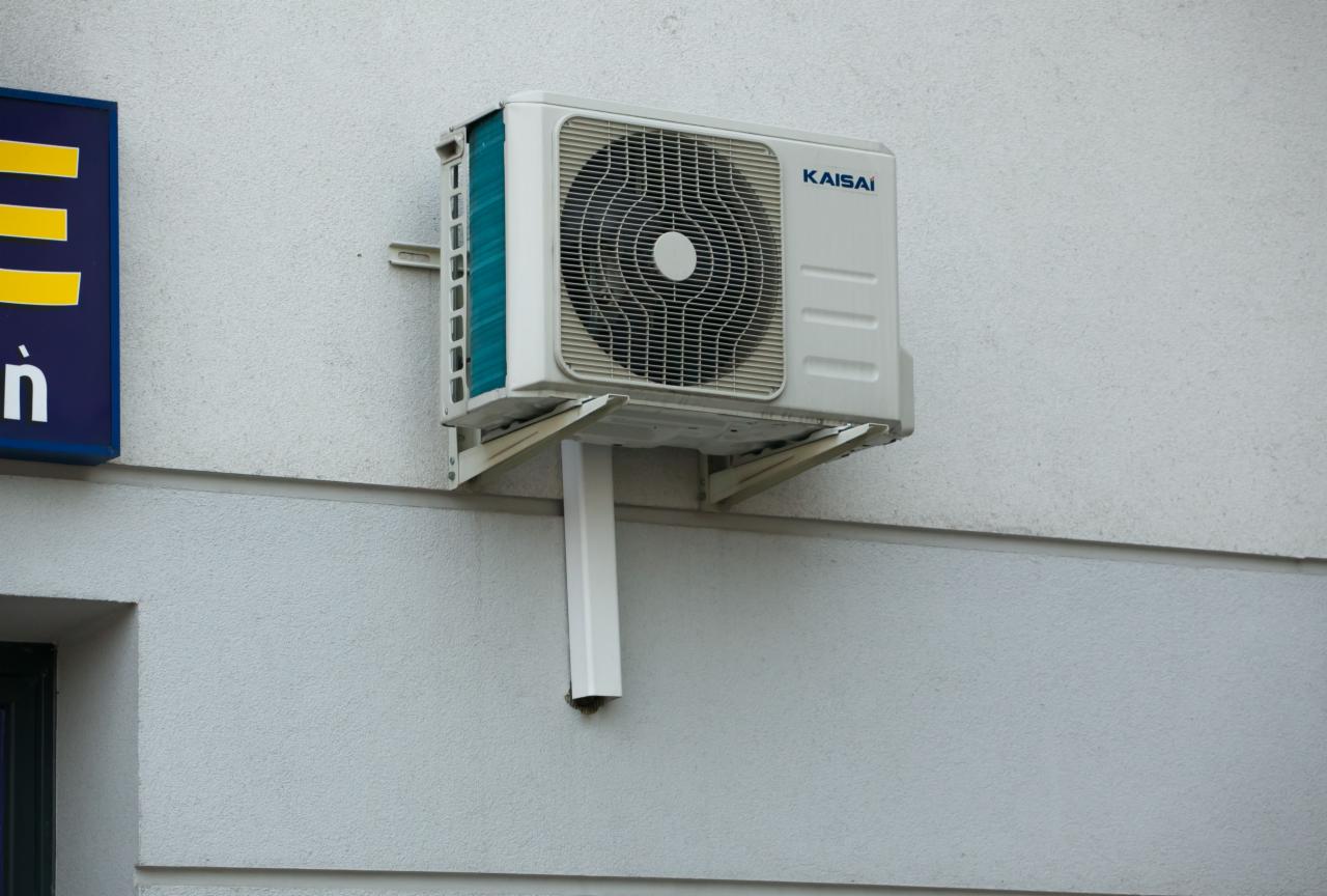Biała jednostka zewnętrzna klimatyzacji marki Kaisai zamontowana na szarej, zewnętrznej ścianie budynku z widoczną rurą odprowadzającą skropliny.