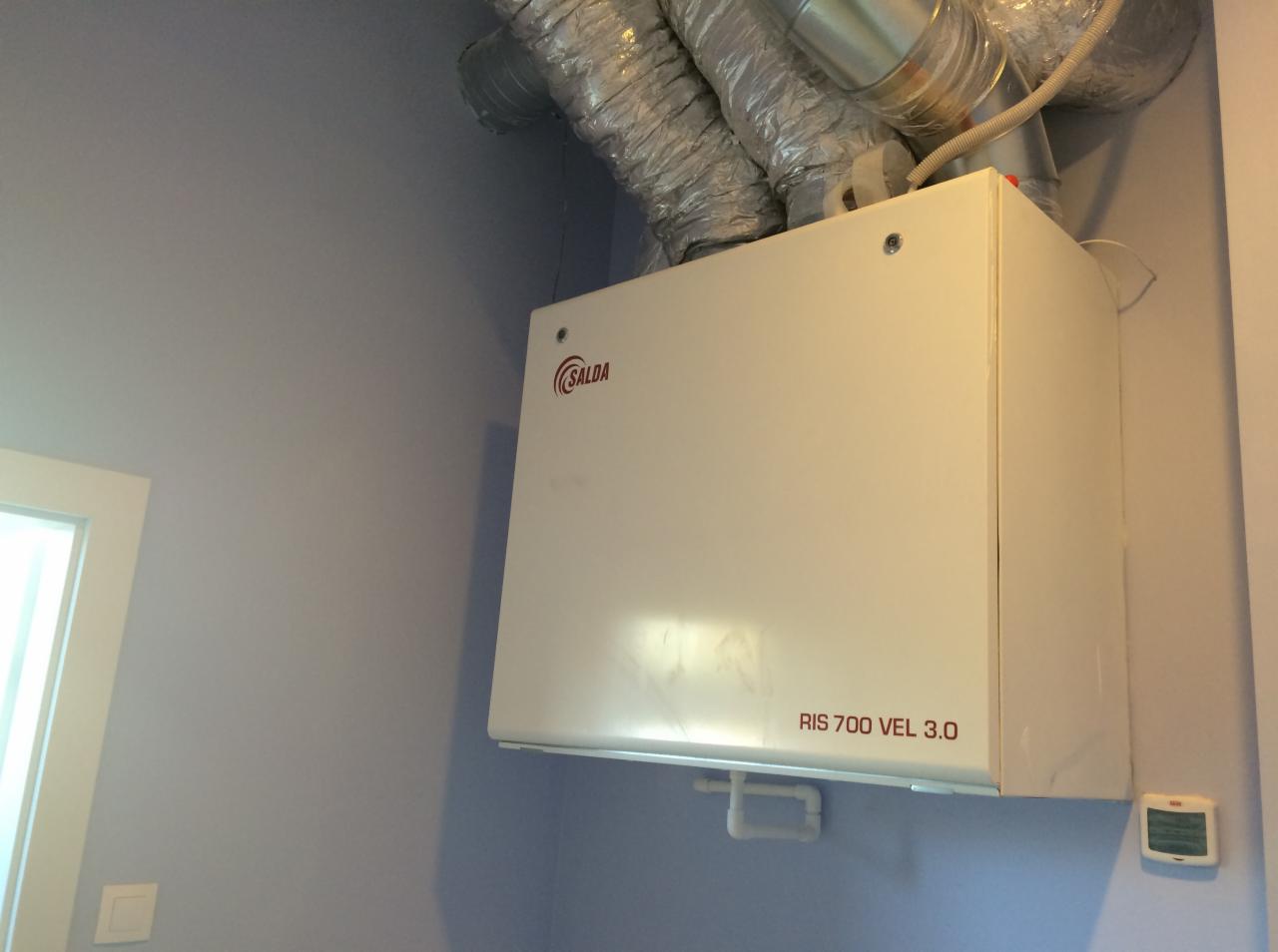 Biała jednostka rekuperacyjna SALDA RIS 700 VEL 3.0 zamontowana na ścianie, z widocznymi przewodami wentylacyjnymi w górnej części i panelem sterowania na ścianie obok.