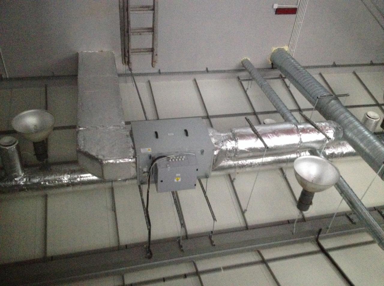 Widok instalacji wentylacyjnej pod sufitem w budynku komercyjnym, z widocznymi przewodami izolowanymi folią aluminiową, centralą wentylacyjną i lampami oświetleniowymi.