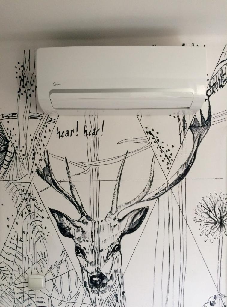 Biały klimatyzator Midea zamontowany na ścianie z czarno-białym rysunkiem jelenia i geometrycznymi wzorami.