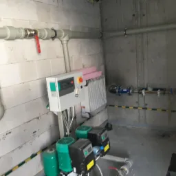 Pomieszczenie techniczne z zainstalowanymi pompami obiegowymi, rurami z zaworem kulowym z czerwoną rączką, centralą sterującą i grzejnikiem panelowym na ścianie z bloczków betonowych.