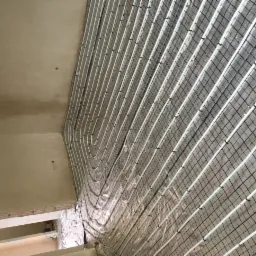 Montaż ogrzewania podłogowego z białymi rurkami na siatce, widok ukośny na surowy strop i ścianę.