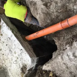 Pracownik w jaskrawej kurtce łączy pomarańczową rurę kanalizacyjną z betonowym zbiornikiem w wykopie ziemnym.