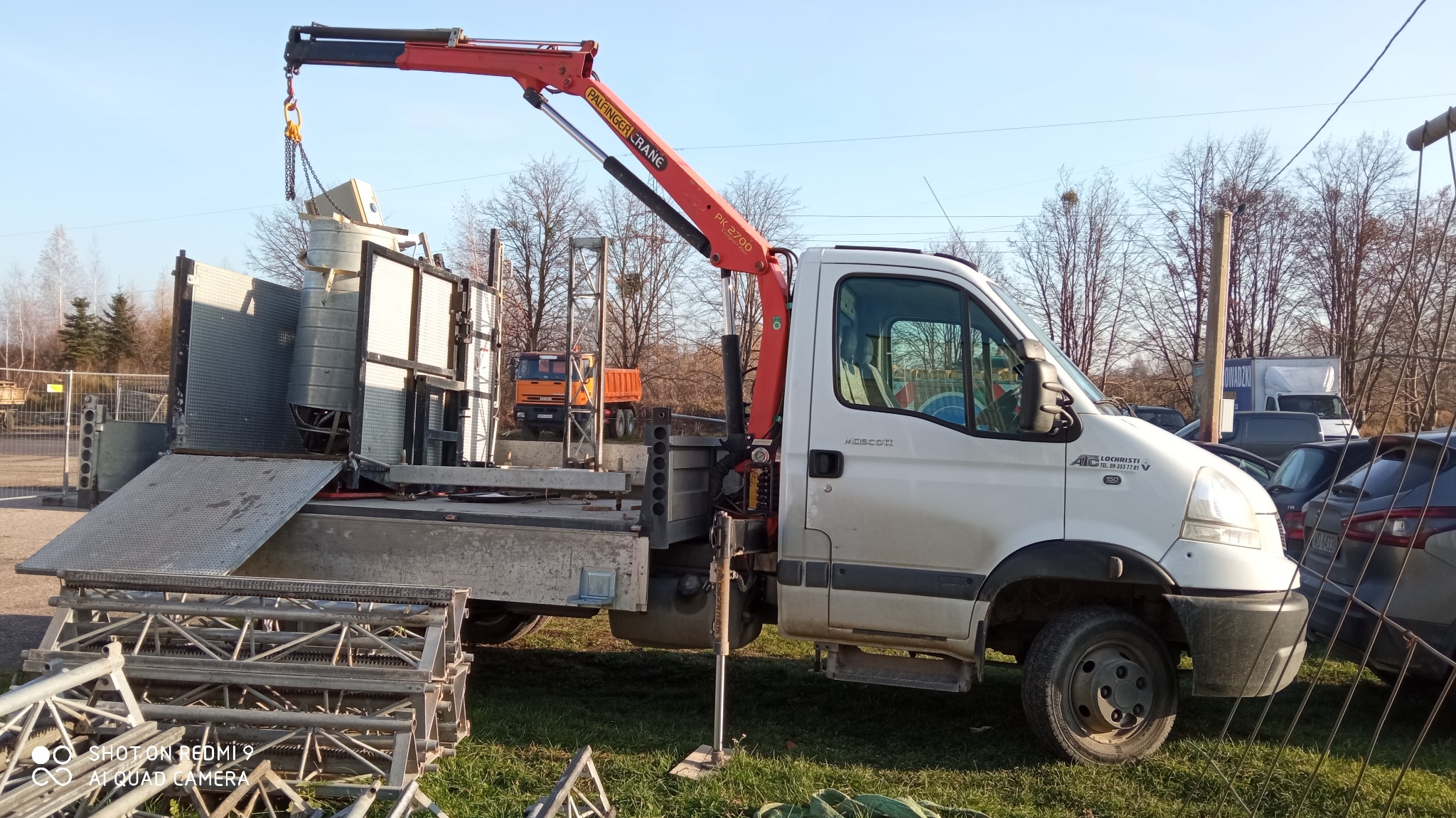 Biały samochód dostawczy z zamontowanym czerwonym dźwigiem Palfinger Crane PK 2700, podnoszący element konstrukcyjny, na trawiastym terenie, w tle inne pojazdy i drzewa.