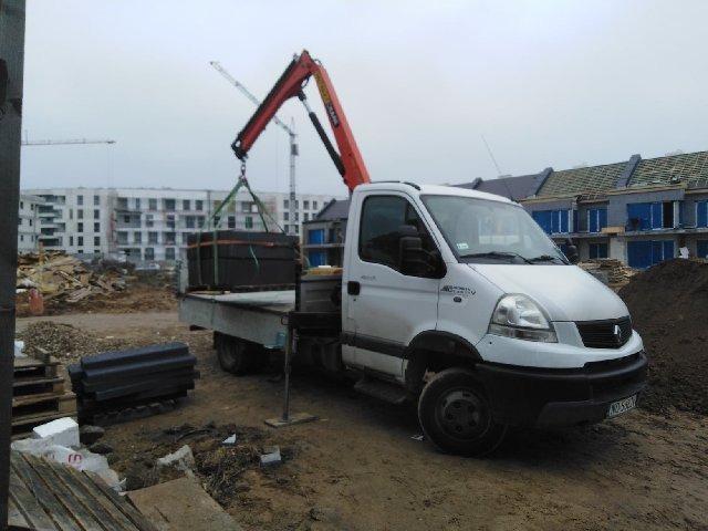 Biały bus z dźwigiem HDS na placu budowy, transportujący czarne elementy konstrukcyjne, w tle budynki w stanie surowym.