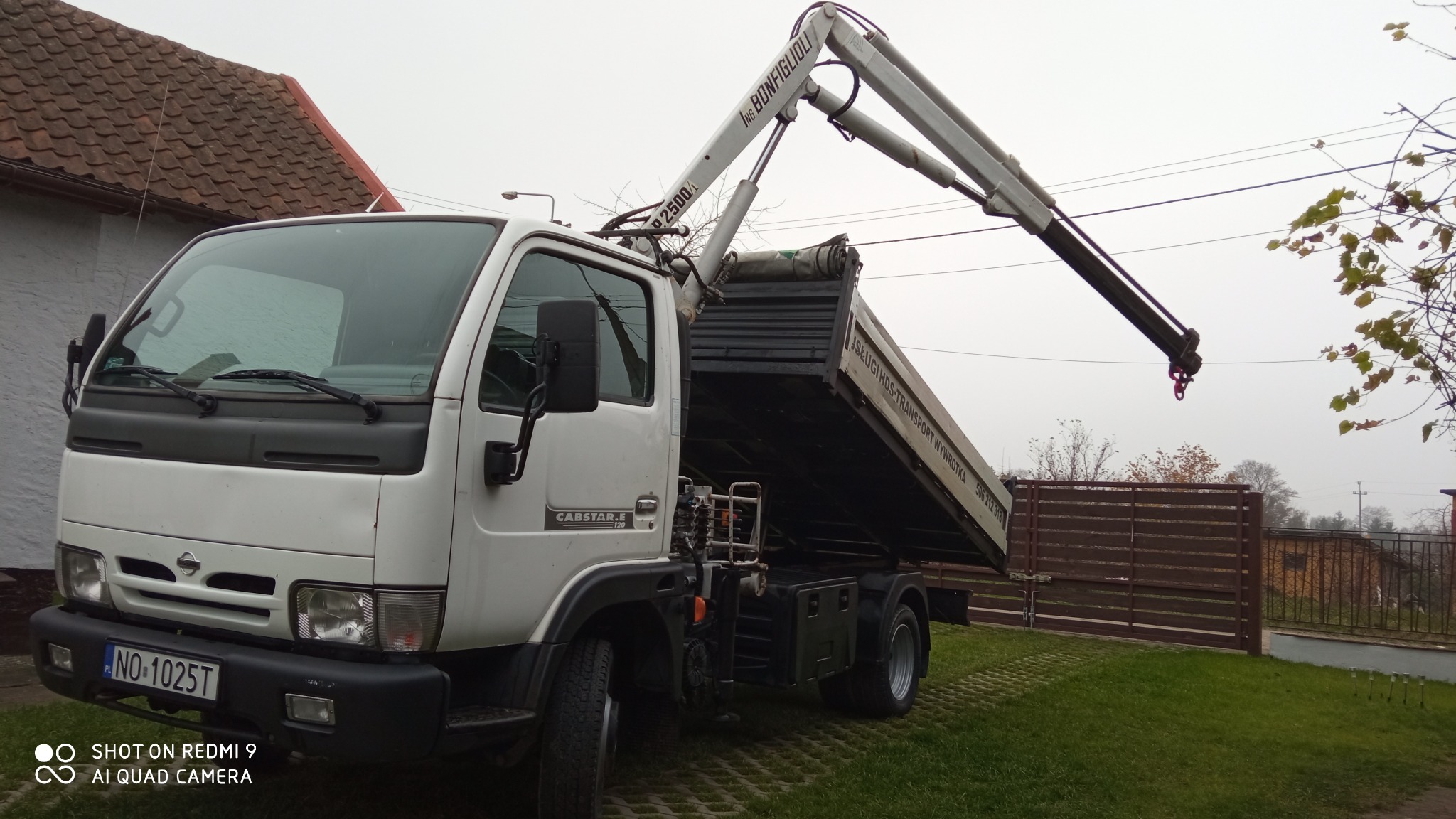 Biały samochód ciężarowy z dźwigiem HDS w Olsztynie, z podniesioną skrzynią ładunkową i wysuniętym ramieniem dźwigu, na tle trawnika i ogrodzenia.