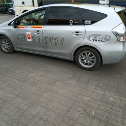 taxi osobowe - Srebrna taksówka ELE Taxi z numerem telefonu i logo iTaxi, zaparkowana na brukowanej nawierzchni. Widok z boku, zarys sylwetki auta.
