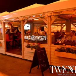 Zewnętrzny ogródek restauracji Twenty 20 Club w Bydgoszczy, widoczne osoby siedzące przy stolikach, drewniana konstrukcja z kwiatami w skrzynkach i tablica z menu.