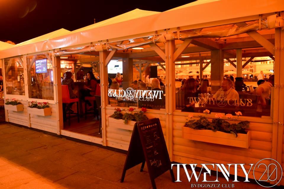 Zewnętrzny ogródek restauracji Twenty 20 Club w Bydgoszczy, widoczne osoby siedzące przy stolikach, drewniana konstrukcja z kwiatami w skrzynkach i tablica z menu.